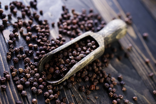 Organic black peppercorns spilling from a rustic wooden scoop onto a dark wooden table, showcasing their natural texture and rich color.